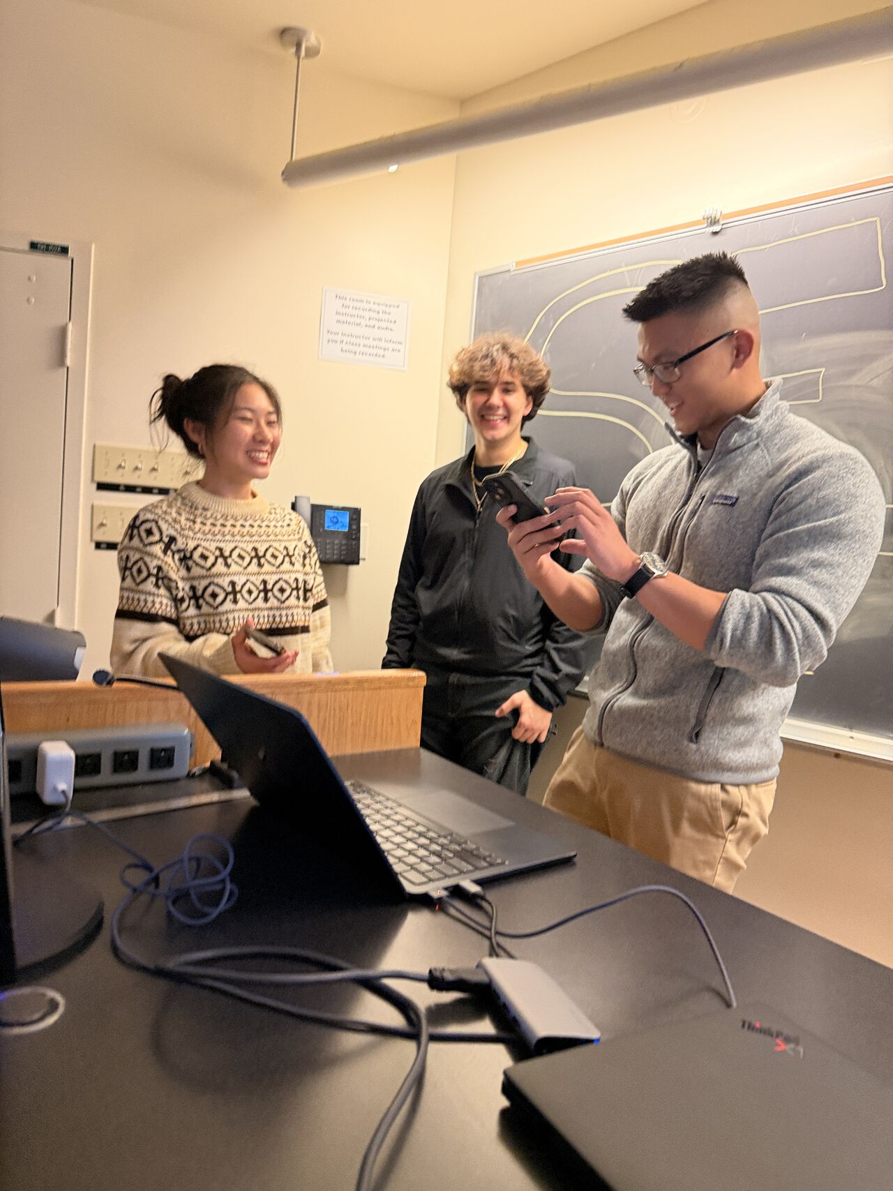 Three students laughing at the front of a lecture room before the joint club meeting begins